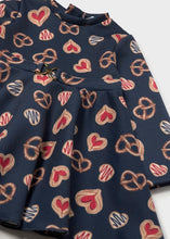 Load image into Gallery viewer, A close-up of a navy blue long-sleeve dress with an allover heart print in red, and beige, featuring a decorative metal ring detail at the waist.
