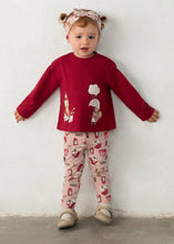Load image into Gallery viewer, A girl toddler stands against a white wall wearing a red long-sleeve top with a whimsical character print, matching pink patterned pants, and a pink patterned headband. Mayoral set available on kidstuff.ie