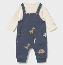 Load image into Gallery viewer, A baby's long-sleeve overall romper in denim-blue colour with a dinosaur pattern, layered over a cream-colored long-sleeve shirt. The overall features adjustable shoulder straps with wooden buttons, front patch pockets, and leg cuffs, available on kidstuff.ie
