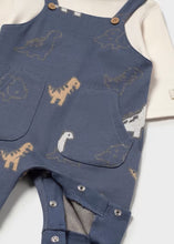 Load image into Gallery viewer, Close-up of a baby's blue denim-style overall romper with a dinosaur pattern, layered over a cream-colored long-sleeve shirt. The overall features front pockets and snap closures.
